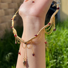 Load image into Gallery viewer, A hand holding up an Intricately hand braided wire wrapped necklace made with Gold Plated Wire lined with red garnet gemstones, Gold Plated Diamond Cut chain festoons with dripping red garnet accent gems, and finished with a gold wire wrapped pear shaped red garnet pendant with swirly accents, against greenery in the background.
