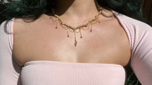 Load image into Gallery viewer, a close up of a woman in a pink dress showcasing an Intricately hand braided wire wrapped necklace made with Gold Plated Wire lined with pink tourmaline gemstones, Gold Plated Diamond Cut chain festoons with dripping pink tourmaline accent gems, and finished with a gold wire wrapped pear shaped pink tourmaline pendant with swirly accents, against greenery in the background.

