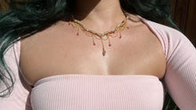 Load image into Gallery viewer, a close up of a woman in a pink dress showcasing an Intricately hand braided wire wrapped necklace made with Gold Plated Wire lined with pink tourmaline gemstones, Gold Plated Diamond Cut chain festoons with dripping pink tourmaline accent gems, and finished with a gold wire wrapped pear shaped pink tourmaline pendant with swirly accents, against greenery in the background.
