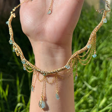 Load image into Gallery viewer, A hand holding up an Intricately hand braided wire wrapped necklace made with Gold Plated Wire lined with blue apatite gemstones, Gold Plated Diamond Cut chain festoons with dripping blue apatite accent gems, and finished with a gold wire wrapped oval shaped blue aquamarine pendant with swirly accents, against greenery in the background.
