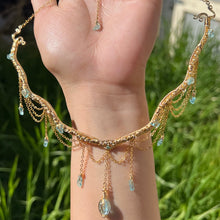 Load image into Gallery viewer, A hand holding up an Intricately hand braided wire wrapped necklace made with Gold Plated Wire lined with blue apatite gemstones, Gold Plated Diamond Cut chain festoons with dripping blue apatite accent gems, and finished with a gold wire wrapped oval shaped blue aquamarine pendant with swirly accents, against greenery in the background.
