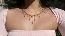 Load image into Gallery viewer, a close up of a woman in a pink dress showcasing an Intricately hand braided wire wrapped necklace made with Gold Plated Wire lined with blue tourmaline gemstones, Gold Plated Diamond Cut chain festoons with dripping blue tourmaline accent gems, and finished with a gold wire wrapped pear shaped blue topaz pendant with swirly accents, against greenery in the background.
