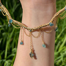 Load image into Gallery viewer, A hand holding up an Intricately hand braided wire wrapped necklace made with Gold Plated Wire lined with blue tourmaline gemstones, Gold Plated Diamond Cut chain festoons with dripping blue tourmaline accent gems, and finished with a gold wire wrapped pear shaped blue topaz pendant with swirly accents, against greenery in the background.
