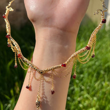Load image into Gallery viewer, A hand holding up an Intricately hand braided wire wrapped necklace made with Gold Plated Wire lined with red garnet gemstones, Gold Plated Diamond Cut chain festoons with dripping red garnet accent gems, and finished with a gold wire wrapped pear shaped red garnet pendant with swirly accents, against greenery in the background.

