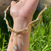 Load image into Gallery viewer, A hand holding up an Intricately hand braided wire wrapped necklace made with Gold Plated Wire lined with blue apatite gemstones, Gold Plated Diamond Cut chain festoons with dripping blue apatite accent gems, and finished with a gold wire wrapped oval shaped blue aquamarine pendant with swirly accents, against greenery in the background.
