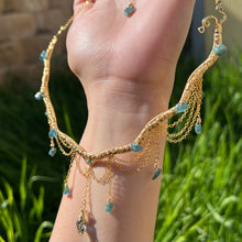 Load image into Gallery viewer, A hand holding up an Intricately hand braided wire wrapped necklace made with Gold Plated Wire lined with blue tourmaline gemstones, Gold Plated Diamond Cut chain festoons with dripping blue tourmaline accent gems, and finished with a gold wire wrapped pear shaped blue topaz pendant with swirly accents, against greenery in the background.
